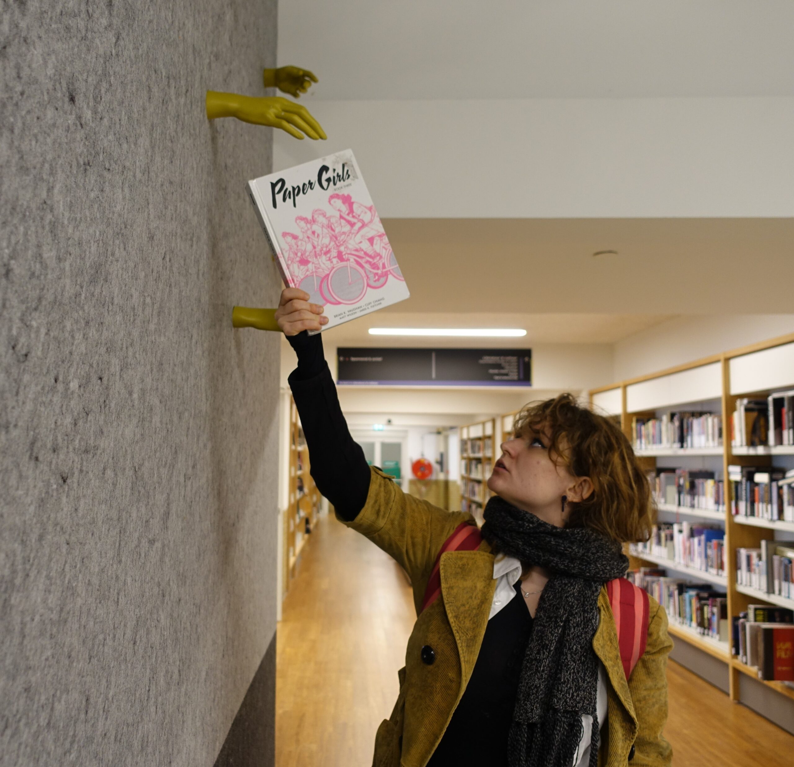 A young person reaches their right hand towards an art installation of a golden hand. The golden hand seems to be handing them a book. The book is white with a pink drawing, and the title of the book, Paper Girls, is written in black. There are book shelves visible in the background. The background is grey felt on the left, and white and honey-coloured on the right.