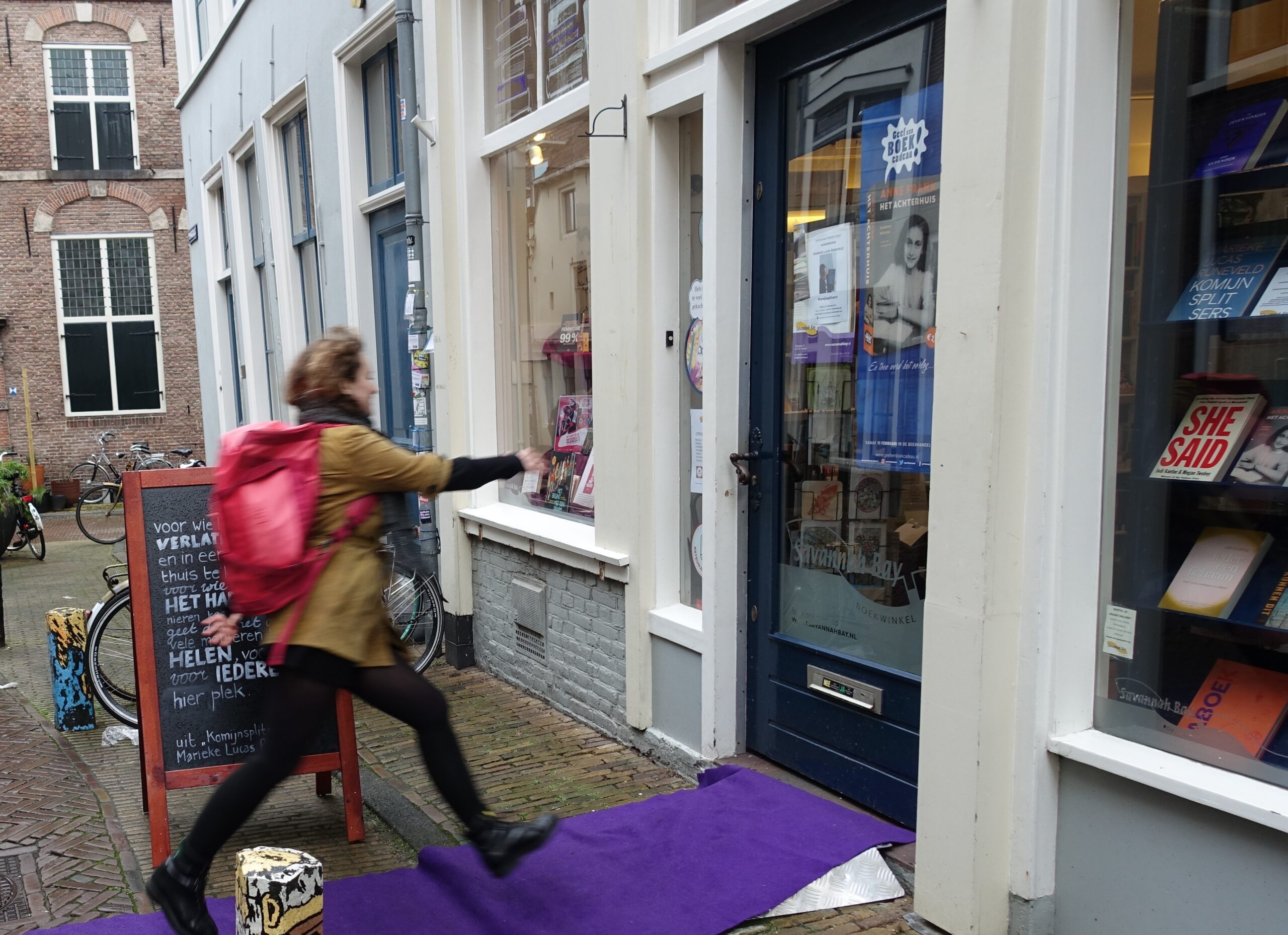 A young person is walking towards the Savannah Bay bookshop. They are shown to be walking at brisk speed and with purpose, just stepping onto a purple carpet, a couple of feet from the door. The door of the bookshop has a wooden, dark blue frame, with glass in the middle. On both side of the door are windows that are displaying books.