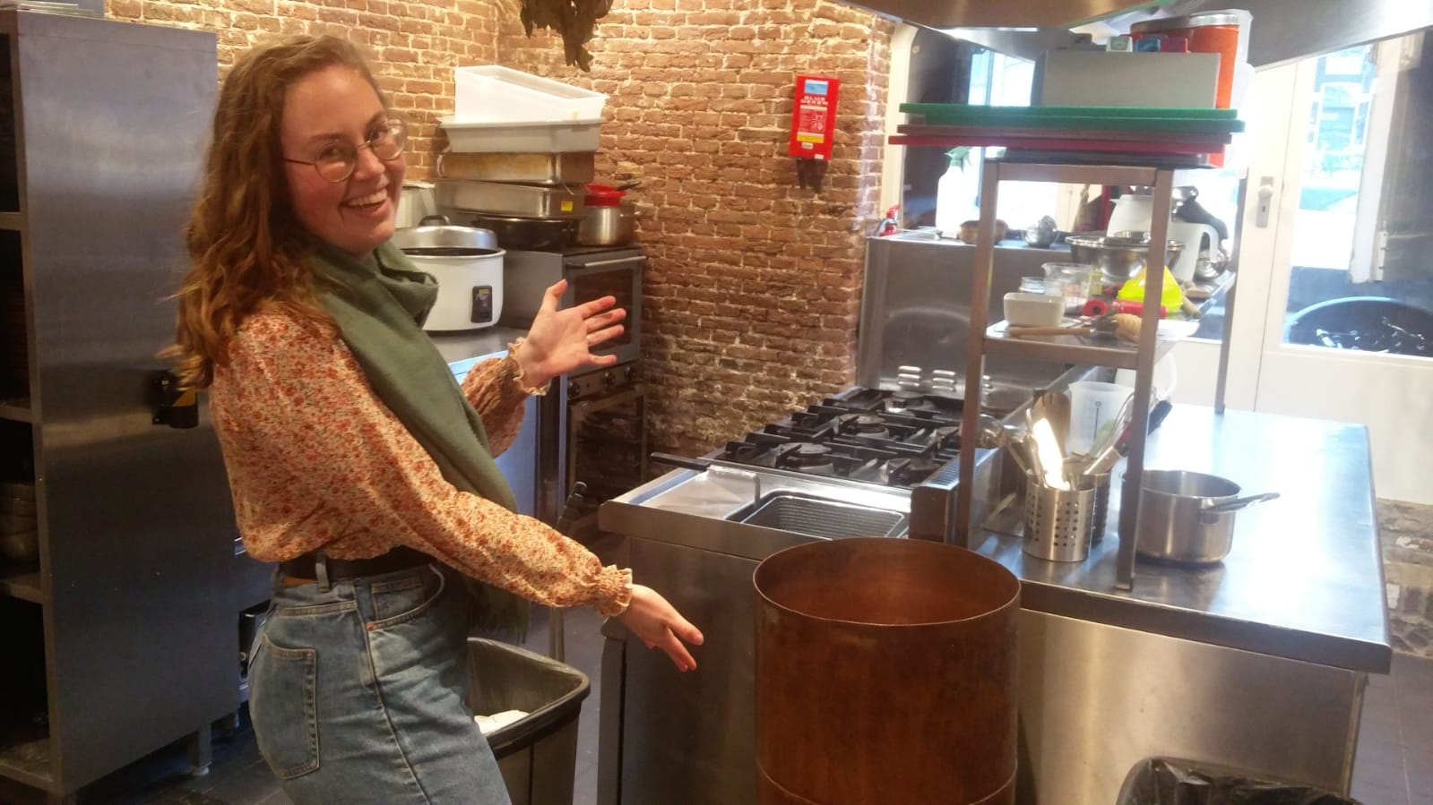 Julia, a young adult with long hair and glasses, with beer brewing equipment, in an industrial sized kitchen.
