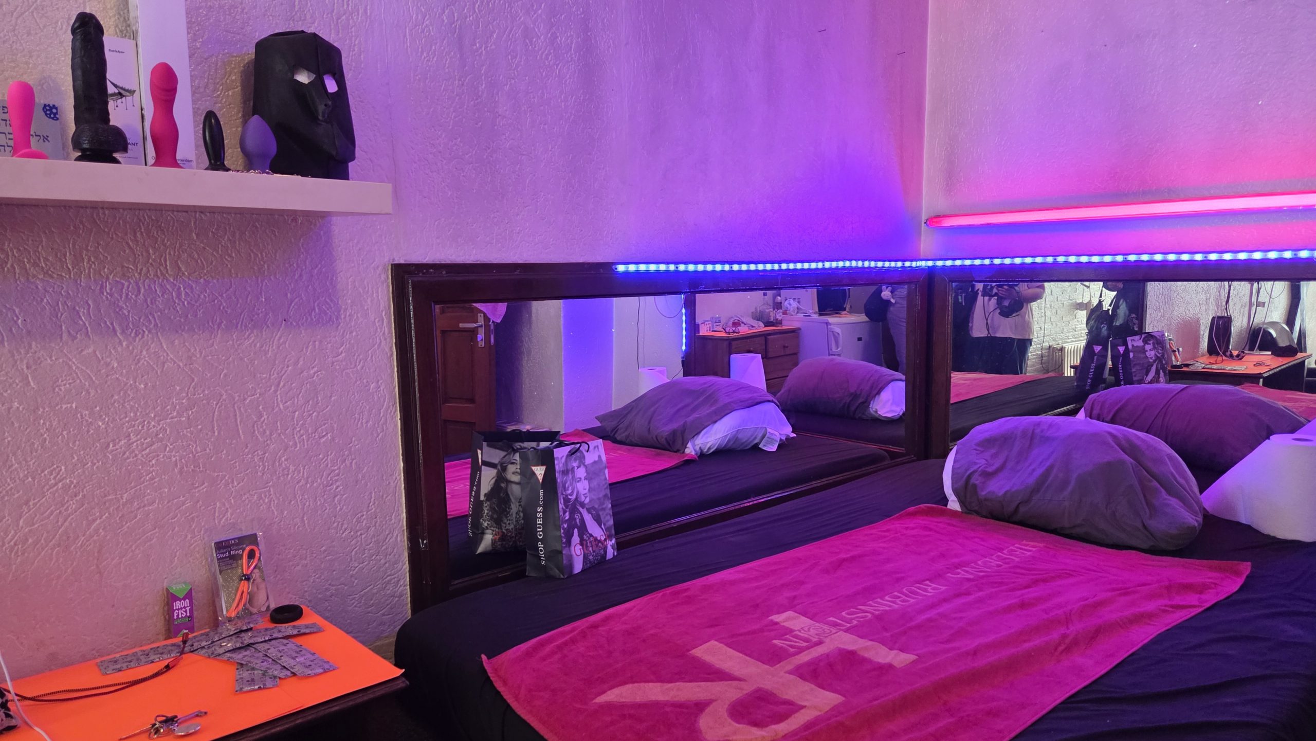 Interior of a red‑light room with a large bed, mirror wall, LED strip lights and a shelf displaying dildos and other sex toys.