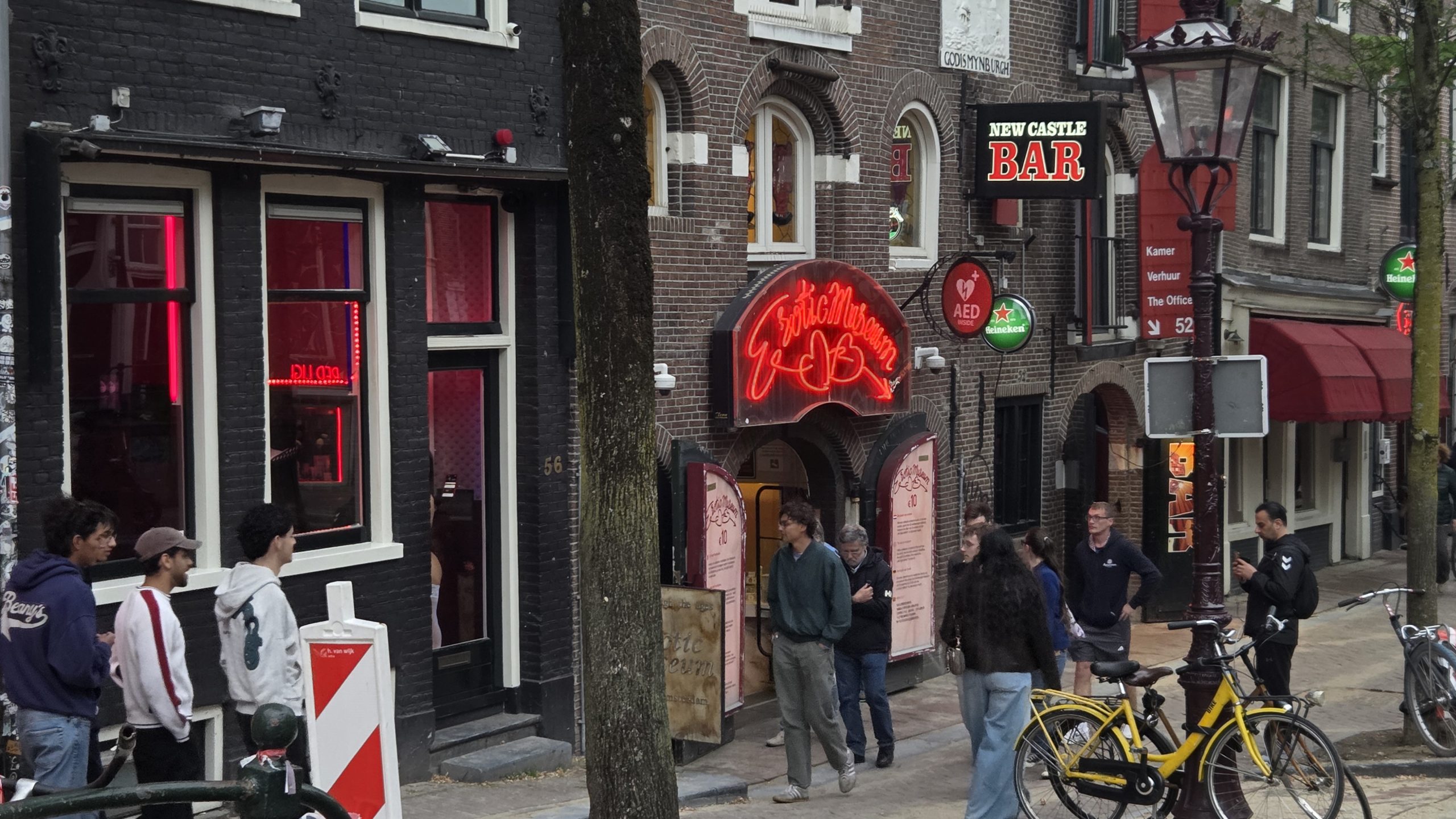Group of people standing in front of the neon sign of the Erotic Museum, next to bars and red‑lit windows in an Amsterdam street.