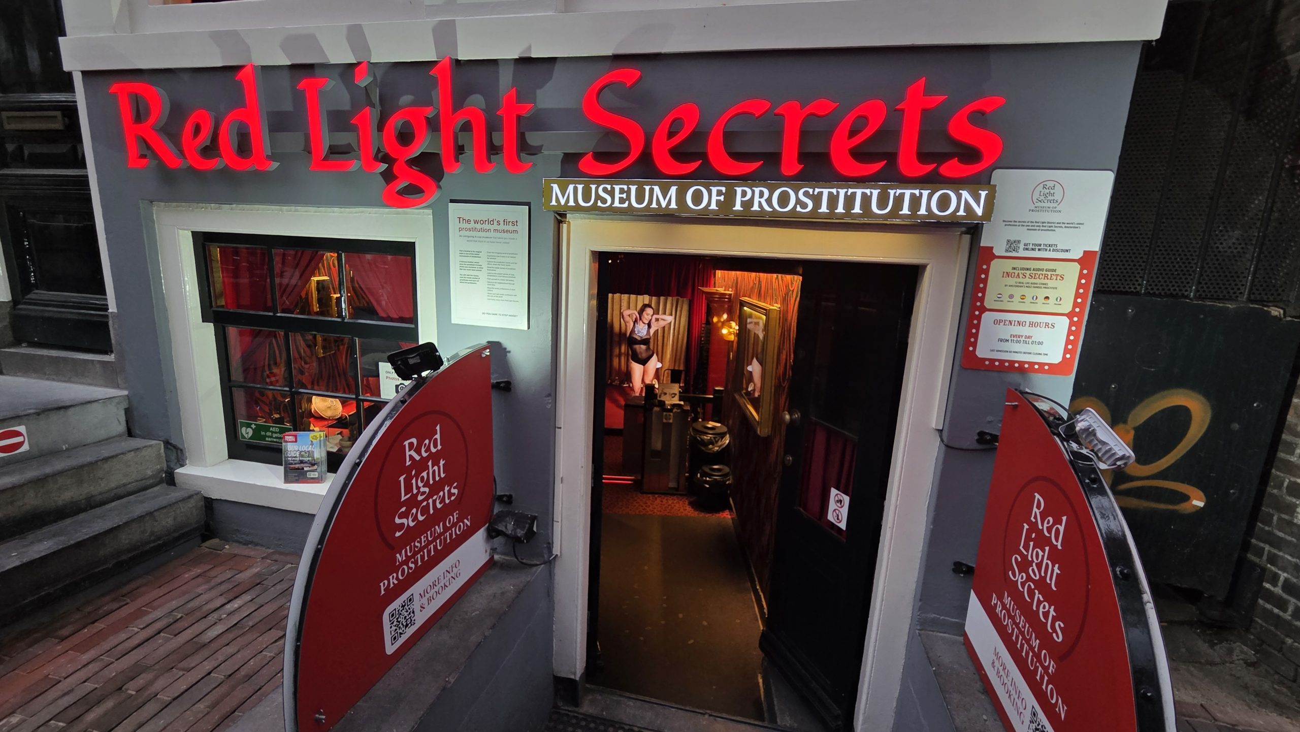 Glowing “Red Light Secrets – Museum of Prostitution” sign above a stairway entrance, looking into a red‑lit interior room.
